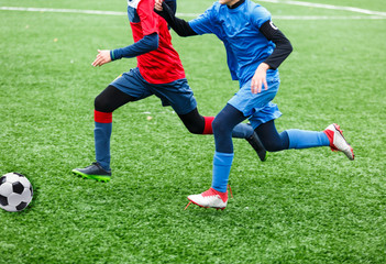 Children are kicking soccer classic white and black ball. Young kids football action. Boys are running after the Ball on green artificial grass. Footballers in blue and red shirts