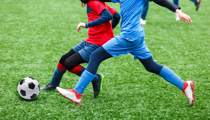 Children are kicking soccer classic white and black ball. Young kids football action. Boys are running after the Ball on green artificial grass. Footballers in blue and red shirts