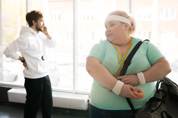Waist up portrait of obese young woman holding donut leaving fitness training, copy space