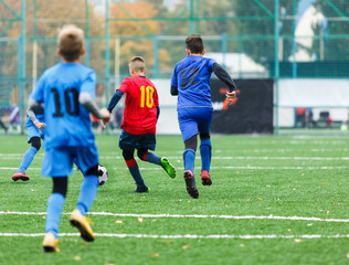 Fototapeta premium Children are kicking soccer classic white and black ball. Young kids football action. Boys are running after the Ball on green artificial grass. Footballers in blue and red shirts