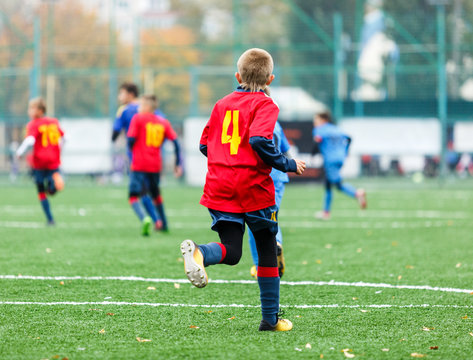 Children Are Kicking Soccer Classic White And Black Ball. Young Kids Football Action. Boys Are Running After The Ball On Green Artificial Grass. Footballers In Blue And Red Shirts