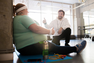 Full length portrait of obese young woman sitting on mat and talking to coach while taking break in...