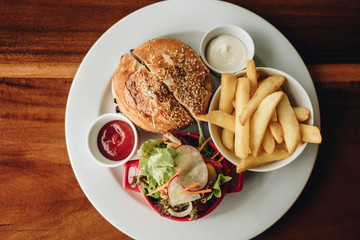 Burger, fresh salad and french fries served on white plate with mayonnaise and tomato sauce on wooden table background