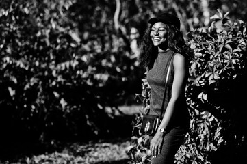 Stylish african american girl in gray tunic, crossbody bag and cap posed at sunny autumn day against red leaves. Africa model woman.