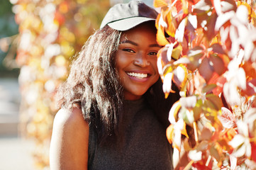 Fototapeta premium Stylish african american girl in cap posed at sunny autumn day against red leaves. Africa model woman.