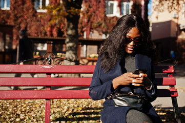 Stylish african american girl in blue coat and sunglasses posed at sunny autumn day at bench and using her mobile phone. Africa model woman with smartphone.