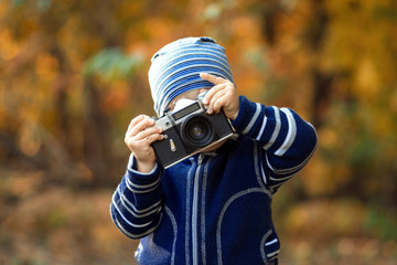 Fototapeta premium Portrait of a little child taking a picture with big vintage photo camera outdoor in autumn forest or park. Brown and golden trees in the background