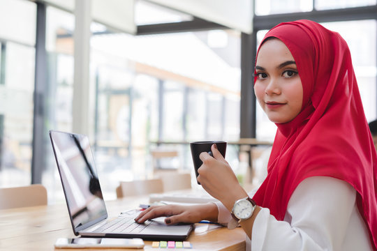 Attractive asian muslim accountant red hijab working with laptop and holding a coffee cup in co-working or coffee shop.Business people working in co-working concept.
