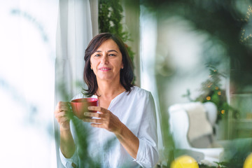 A senior woman with a cup standing by the window at home at Christmas time.