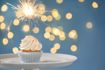 Delicious birthday cupcake with sparkler on stand against blurred lights
