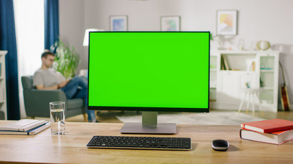 Modern Personal Computer with Green Mock-up Screen Display Standing on the Desk. In the Cozy and Light Living Room Man Relaxes on a Chair Reading a Book.
