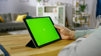 Over the Shoulder Shot of a Man Holding and Watching Green Mock-up Screen Digital Tablet Computer While Sitting at the Desk. In the Background Cozy Living Room.