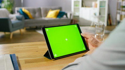 Man Holding and Watching Green Mock-up Screen Digital Tablet Computer While Sitting at the Desk. Man Watching Videos or Browsing Through the Internet. In the Background Cozy Living Room.