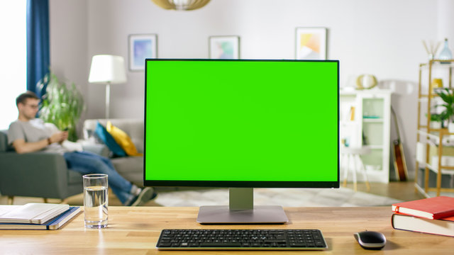 Modern Personal Computer with Green Mock-up Screen Display Standing on the Desk in the Cozy Living Room. A Man with Mobile Phone Relaxes in His Chair.