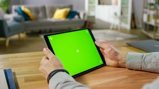 Man Holding and Watching Green Mock-up Screen Digital Tablet Computer While Sitting at the Desk. Man Watching Videos or Browsing Through the Internet. In the Background Cozy Living Room.