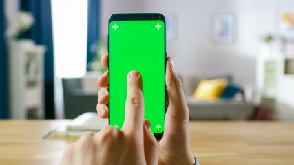 Close-up of a Man Holding Green Mock-up Screen Smartphone and Using Touchscreen Gestures. Touching Mobile Phone Chroma Key Screen. In the Background Cozy Homely Atmosphere.