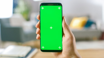 Close-up of a Man's Hand Holding Green Mock-up Screen Smartphone. Modern Mobile Phone. In the Background Cozy Living Room or Home Office.