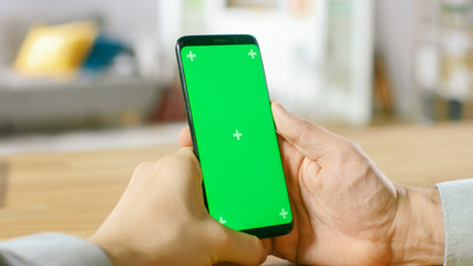 Close-up of a Man Holding Green Mock-up Screen Smartphone in His Hands. Using Wireless Device to Browse Through Internet. In the Background Cozy Living Room or Home Office.