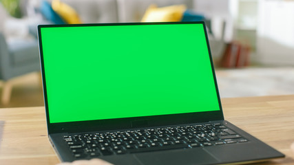 Man Uses Laptop with Green Mock-up Screen While Sitting at the Desk in His Cozy Living Room.