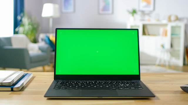 Modern Laptop with Green Mock-up Screen Display Standing on the Desk in the Cozy Living Room. Man with Mobile Phone Walks Through His Flat.
