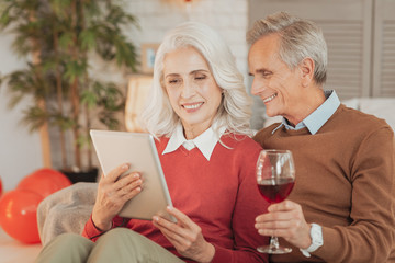 Pleasant pastime. Pleased senior spouses resting in living room with the glass of wine while excited woman showing her husband eyecatching photos on tablet