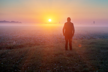 A man stands in the field early in the morning and looks at the sunrise