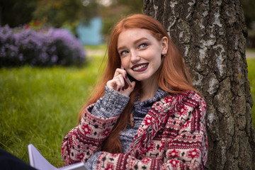 Young smiling woman with freckles and dark lipstick reading book outdoors in park sit under the big tree on grass using mobile phone talking looking aside bite lip.