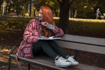 Redhead young cheerful woman with freckles and dark lipstick reading book outdoors in park sit on bench looking aside.