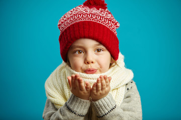 portrait of kid girl in winter red hat, folded her hands together and blowing on camera.