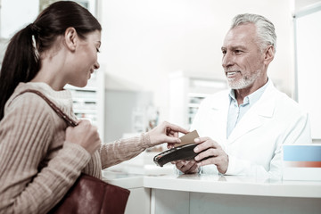 Making payment. Dark-eyed bearded pharmacist wearing white coat holding pin pad while client paying for order by card