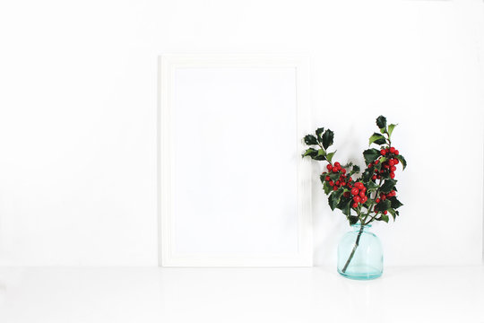 Vertical white blank wooden frame mockup. Holly berry branches in blue glass vase on white table. Styled stock feminine photography. Home decor. Christmas winter concept.