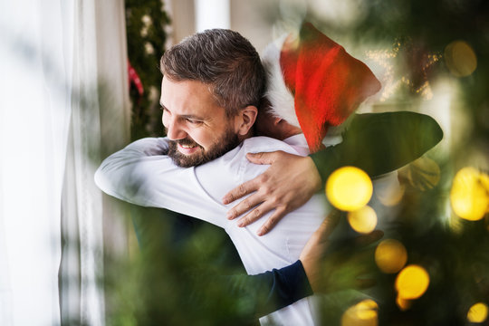 A senior father with a Santa hat and adult son standing by the window, hugging. - Powered by Adobe