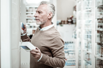 Naklejka na ściany i meble Curious man. Bearded mature grey-haired man feeling curious choosing between two pills standing in pharmacy store
