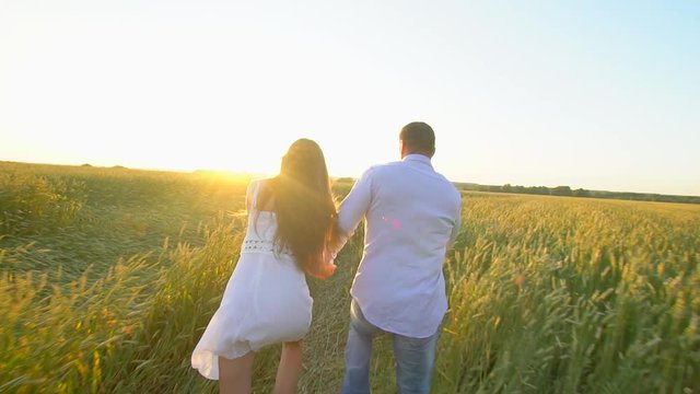Happy young couple holding hands and runing through wheat field at summer sunset, having fun outdoors. Countryside, summer time. Man and woman, happy family in love. Nature. Sun flare Slow motion