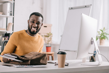 smiling handsome african american designer in orange sweater taking notebooks from briefcase in office