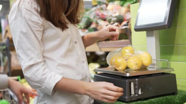 A woman in a white shirt puts a scale on a scale with tangerines in a store. She takes the label and sticks it onto the bag. Buying fruit. Self catering