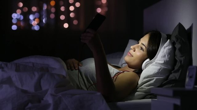 Happy teen with headphones browsing media in a smart phone lying on a bed at home in the night