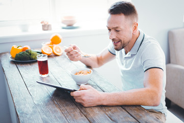 Nutritious meal. Delighted nice man holding a tablet while having his breakfast
