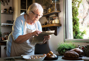 Elderly woman following a cookbook in the kitchen - obrazy, fototapety, plakaty