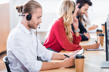 selective focus of call center operators in headsets with notebooks working at workplace in office