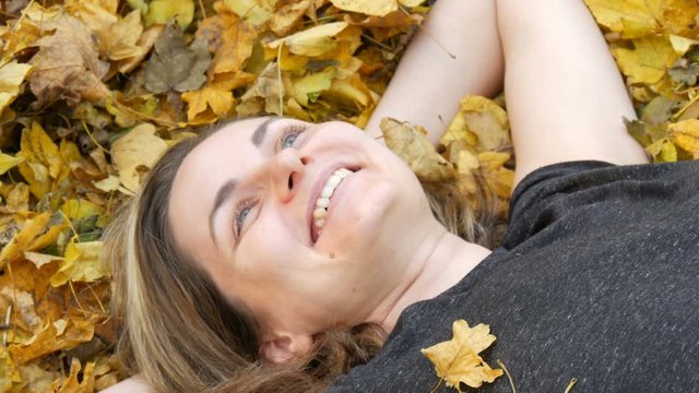 Portrait of the face of beautiful young girl without makeup who lies in the autumn yellow foliage and enjoys life. Dry autumn leaves fall on the face of a laughing woman