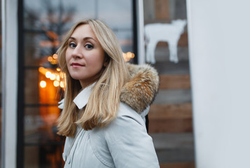 Attractive, charming smiling young woman in a white coat with fur hood walking in the winter street. Background blurry lights. The holiday mood.