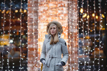 Winter portrait of happy young woman walking in snowy city decorated for Christmas and New Year holidays, wearing in a white coat with fur hood. Christmas lights on background. Black and white photo.