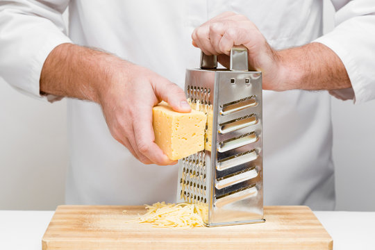 Chef's hands grating piece of cheese with steel grater on the wooden board in professional kitchen. Preparing for italian pizza, lasagna or snack. Front view.