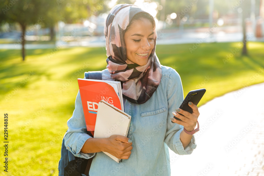 Wall mural happy muslim young woman walking outdoors holding books using mobile phone chatting.