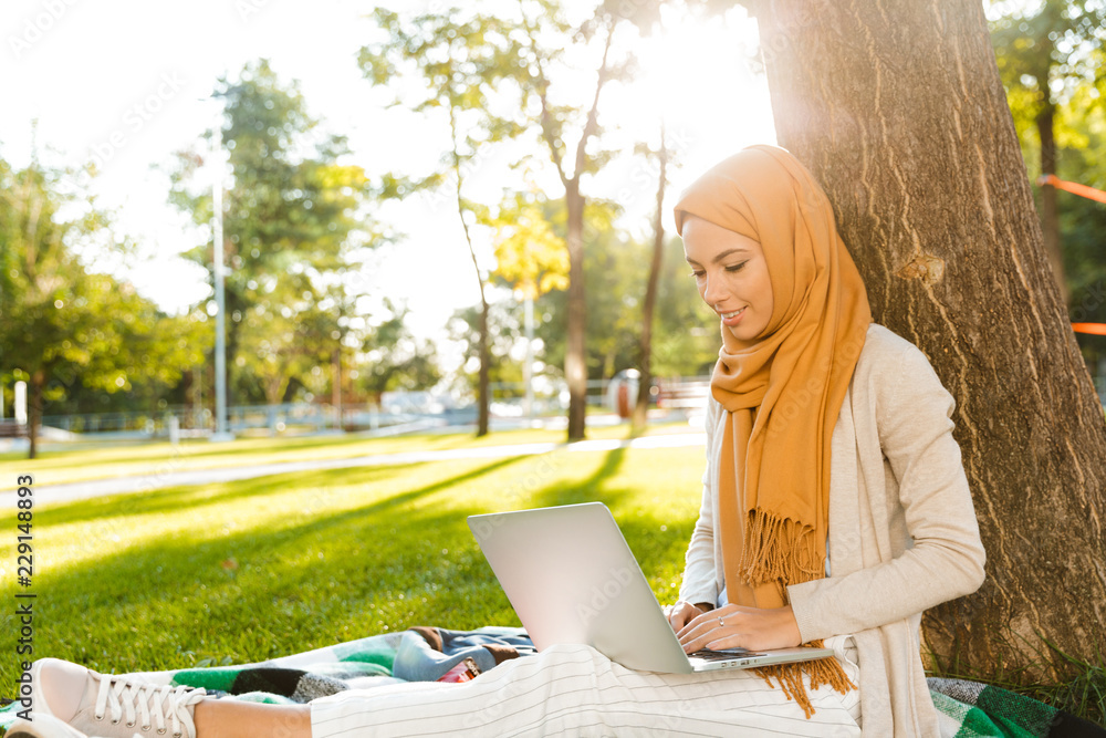 Wall mural happy muslim young woman sitting outdoors in park using laptop computer.