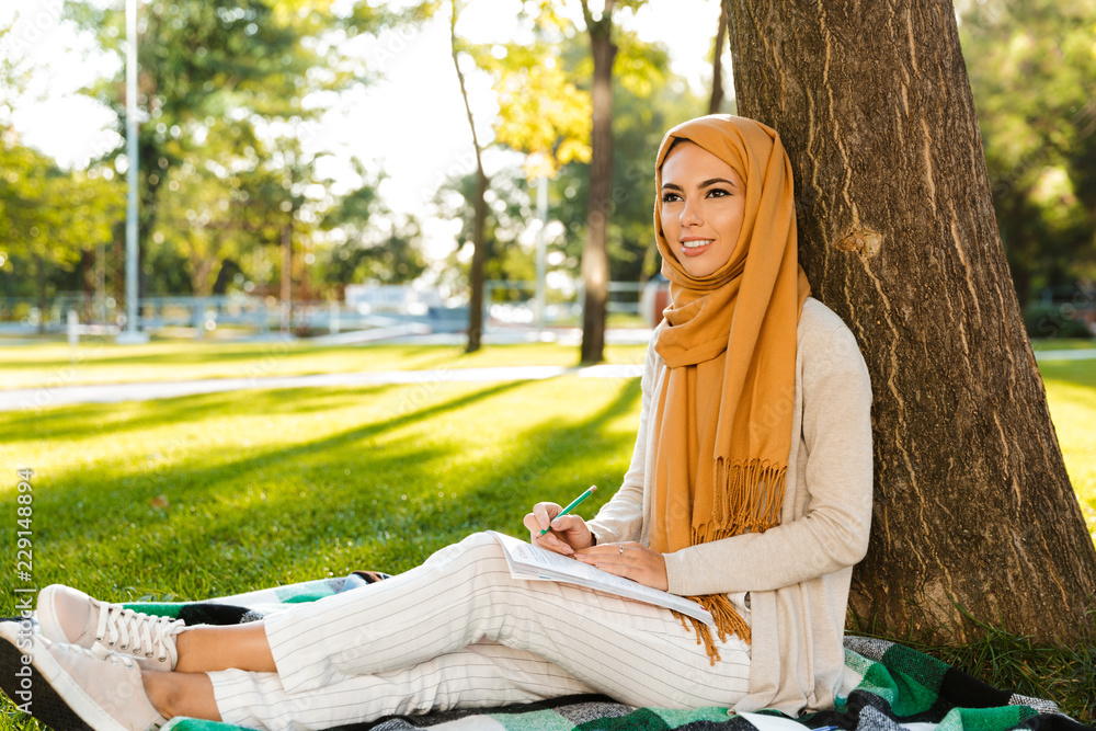 Wall mural happy muslim young woman sitting outdoors in park writing notes.