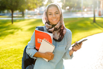Happy muslim young woman walking outdoors holding books using mobile phone chatting.