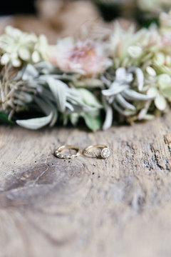 Artisanal designed wedding and engagement ring on a rugged wooden surface with a flower wreath in the background.
