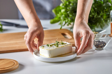 The hands of the girl hold a plate with soft cheese with green sprouts on a white table with dill and parsley, copy space for text. Healthy food